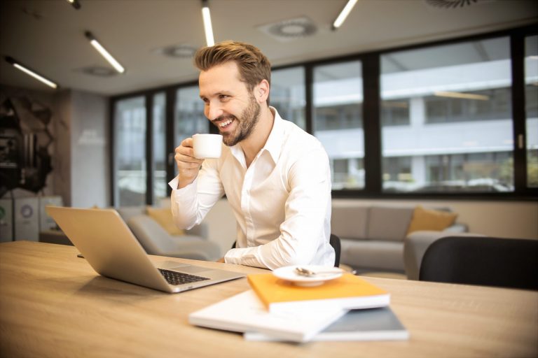 Man holding coffee cup near his mouth smiling with his laptop and some books on the table neat him.