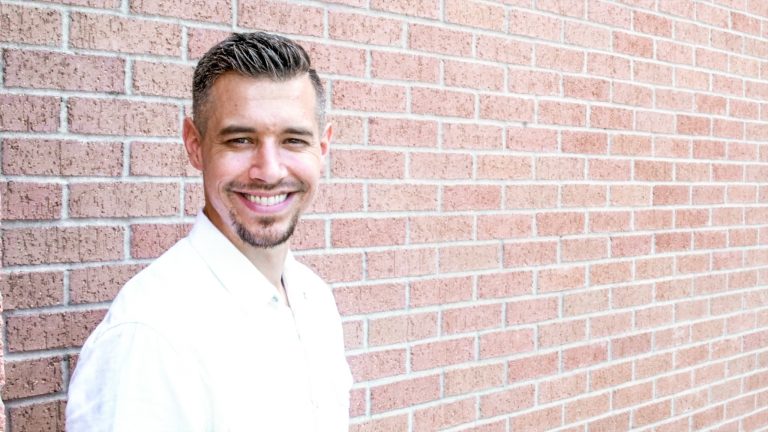 Man in white shirt smiling and standing in front of a brick wall